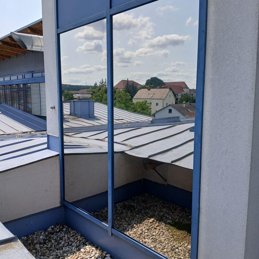Ein Blick aus einem Dachbodenfenster mit Kies auf dem Boden und einem reflektierenden Fenster, das die umliegenden Gebäude zeigt.