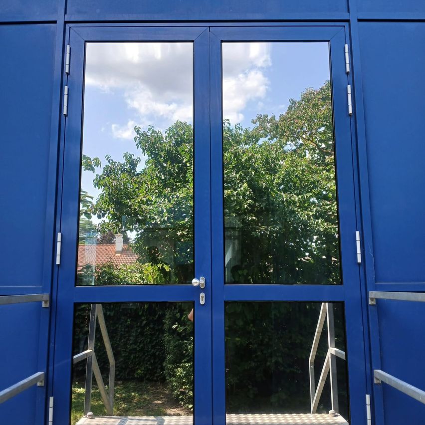 Blaue Doppeltüren mit Glasscheiben reflektieren ein Haus und Grünpflanzen. Metallhandläufe auf beiden Seiten.