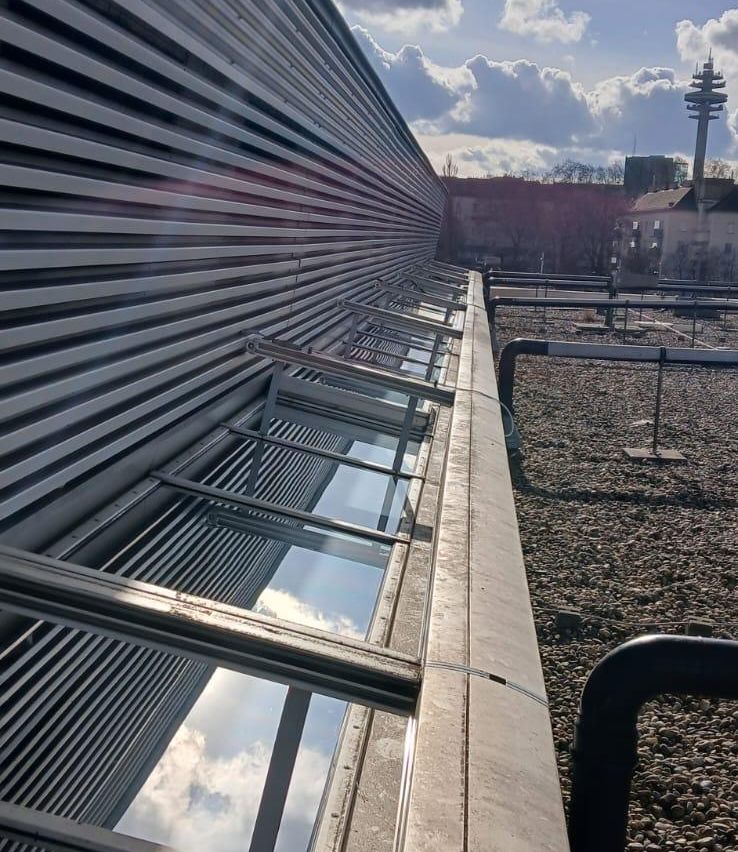 Ein Dach mit Metallgeländern, Glasscheiben und einem Kiesboden. In der Ferne befinden sich Gebäude und Bäume.