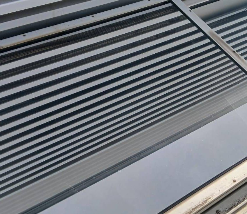 Nahaufnahme eines Metallgitters mit horizontalen Streifen, möglicherweise Teil eines Belüftungssystems. Das Gitter besteht aus Metall mit einer reflektierenden Oberfläche und hat eine kleine Schraube oben links.