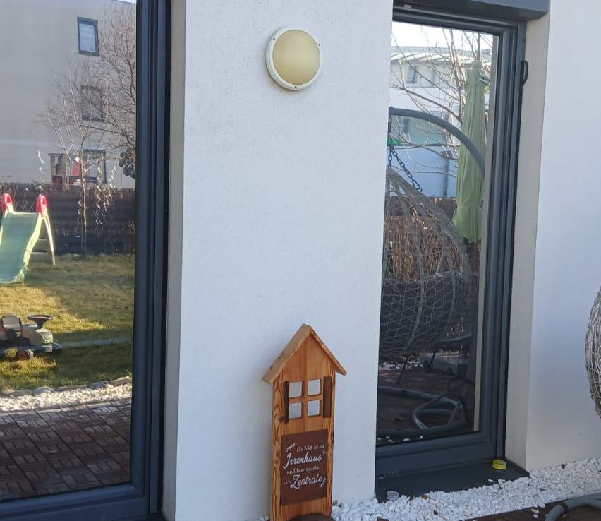 Ein kleines Holzhaus mit einer Tafel steht auf einer Terrasse. Eine Wandlampe ist an einer weißen Wand montiert. Eine Glastür ist offen, und ein Stuhl ist drinnen. Ein Rasen ist vor der Glastür.