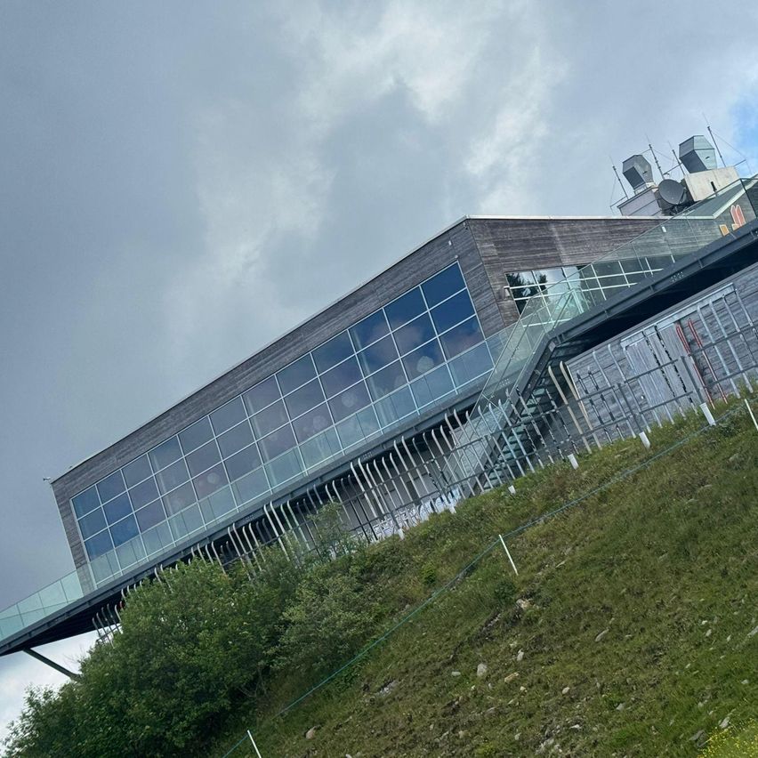 Ein modernes Gebäude mit Glaswänden und einer Glasbrücke auf einem Hügel unter einem bewölkten Himmel.