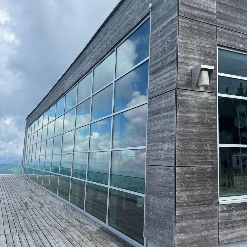 Ein modernes Gebäude mit großen Glasfenstern und einer hölzernen Terrasse, die den Himmel und Wolken reflektiert.