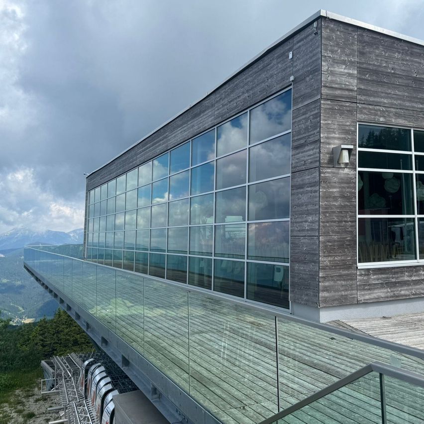 Ein modernes Gebäude mit Glaswänden und einem Balkon, mit Blick auf eine Berglandschaft unter einem bewölkten Himmel.