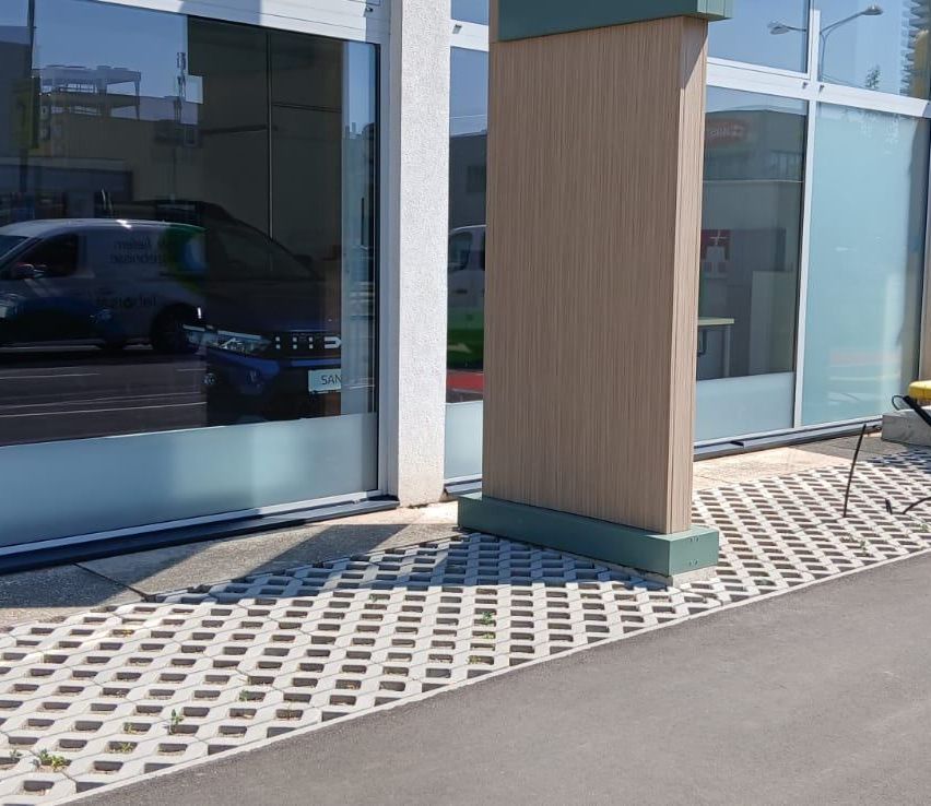 Ein moderner Gebäudeeingang mit einer Glastür, einem hölzernen Pfeiler und einem Bürgersteig mit sechseckigen Fliesen. Ein Auto spiegelt sich in der Glastür.