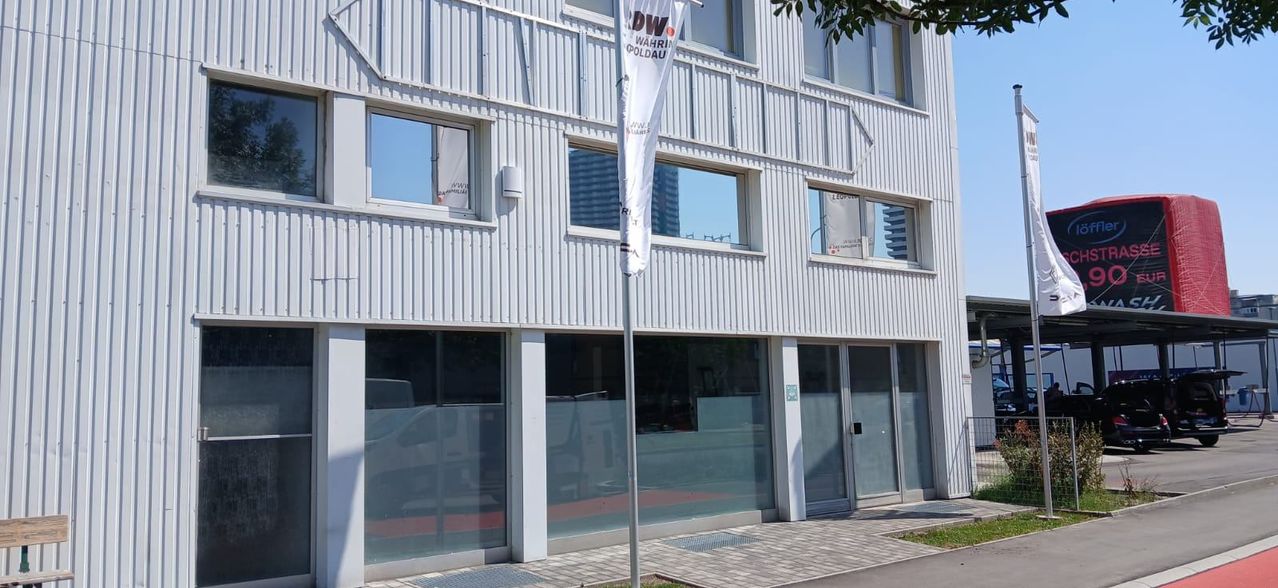 Ein modernes Bürogebäude mit großen Glasfenstern und einer weißen Fahne mit dem Text 'DW' auf einem Pfosten davor.