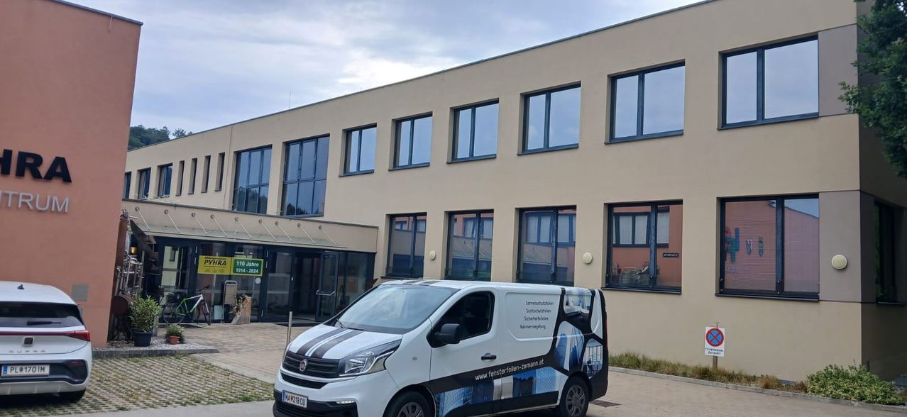 Ein weißer Van mit schwarzen und weißen Streifen ist vor einem Gebäude mit Glasfenstern geparkt. Das Schild des Gebäudes zeigt '10 Jahre 14-2024'. Der Van hat eine Website-Adresse an seiner Seite.