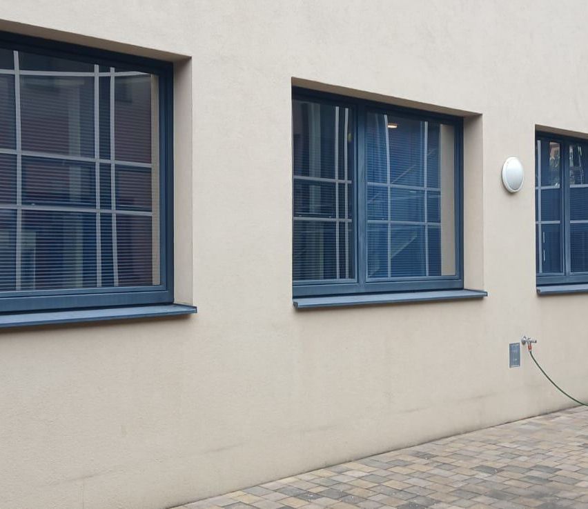 Drei Fenster mit blauen Rahmen an einer beigen Wand. Die Fenster haben Jalousien. Die Wand hat eine weiße Lampe und einen Ziegelboden.