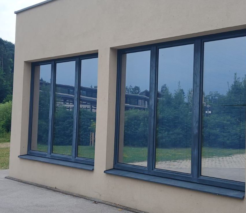 Zwei große Fenster mit blauen Rahmen an einem beigen Gebäude, reflektieren Bäume und ein Gebäude in der Ferne.