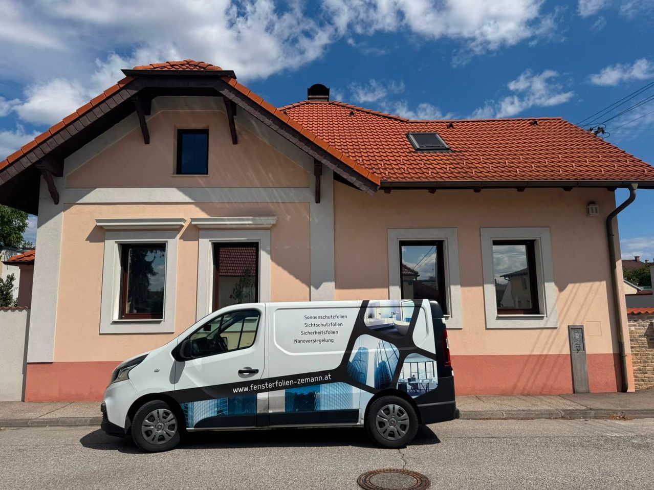 A white van with a black and blue design is parked in front of a house with a red roof and white trim. The van has a website address on its side.