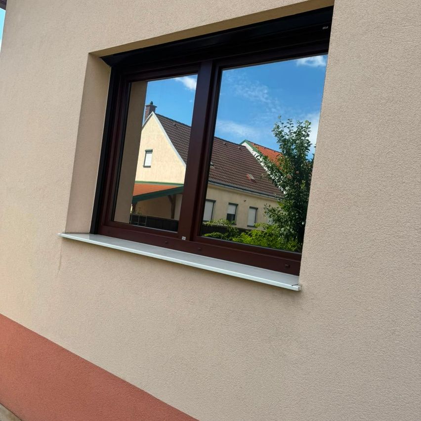 A window with brown frames reflects a house and a tree against a blue sky.