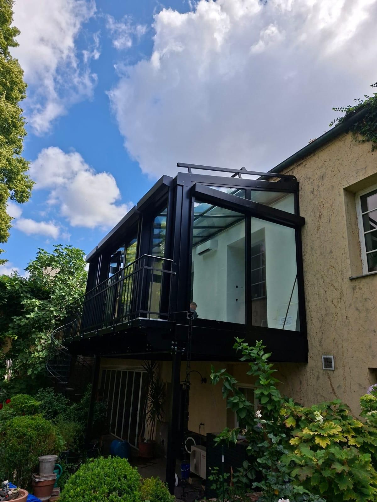 A modern glass balcony with a black frame attached to a beige building, surrounded by greenery under a partly cloudy sky.