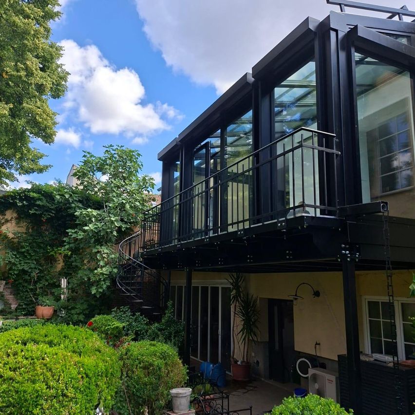 A modern house with a glass balcony, spiral staircase, and lush garden under a partly cloudy sky.