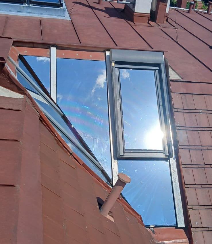 A close-up view of a copper-colored roof with a window open, showing a rainbow reflection on the glass and a clear blue sky with clouds.