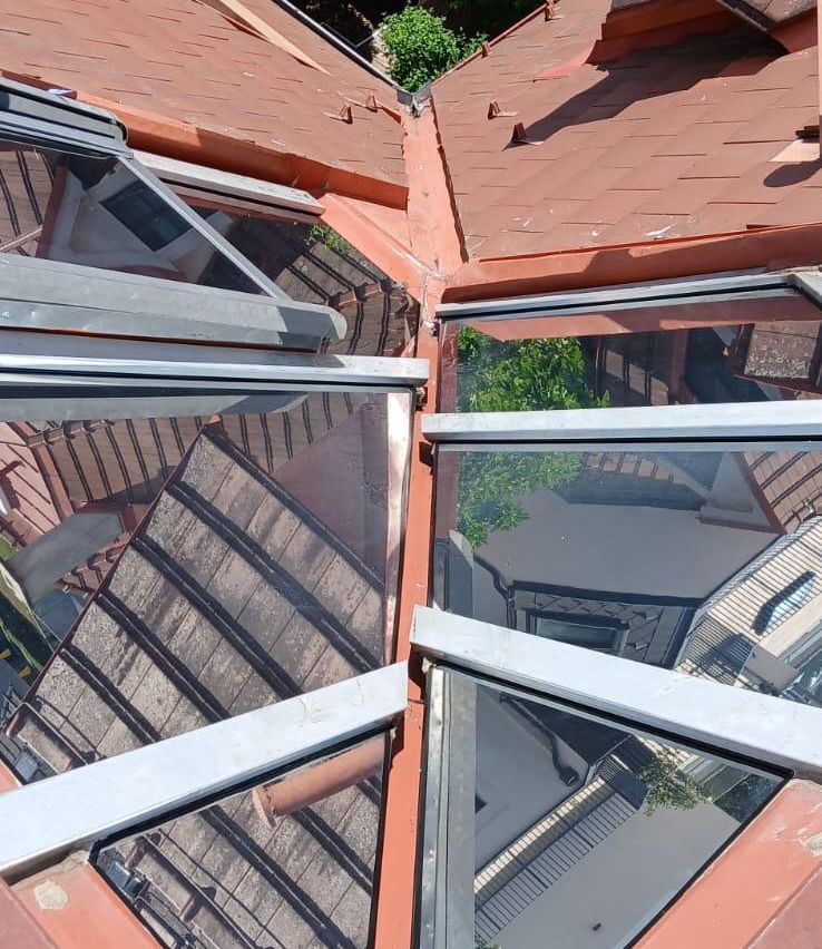 A top-down view of a building's roof with glass panels, metal frames, and a brown roof. The glass panels reflect the surrounding environment, including trees and other buildings.