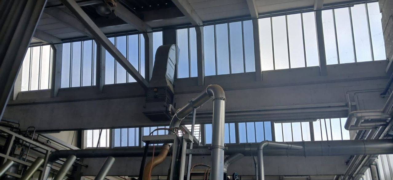 An industrial facility with metal pipes and machinery, large windows allow natural light to enter, casting shadows on the floor.