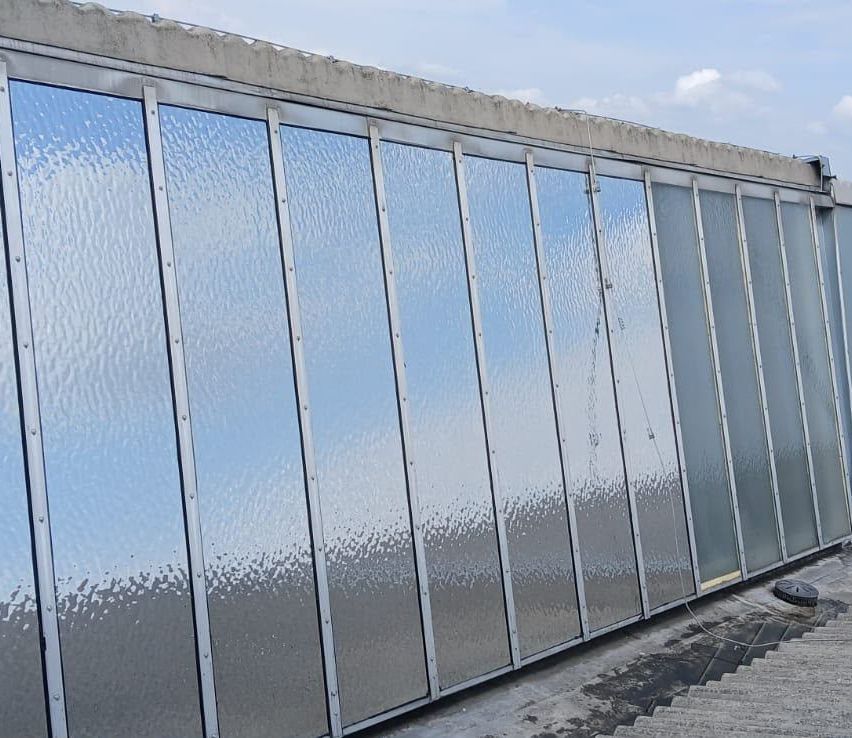 A close-up view of a modern building's exterior wall, featuring multiple large, reflective glass panels with a textured surface. The panels are separated by metal frames and the wall appears to be part of a larger structure.