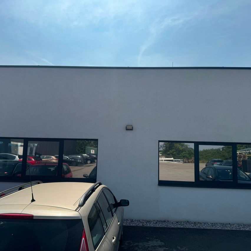 A beige car is parked in front of a white building with large windows, reflecting a parking lot with several cars and trees.