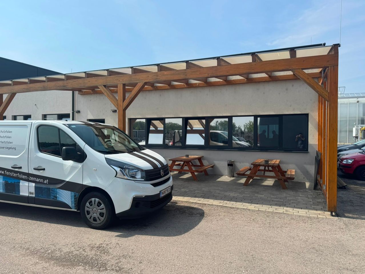 A white van with a logo is parked in front of a building with a wooden pergola. Two wooden picnic tables are on the side.