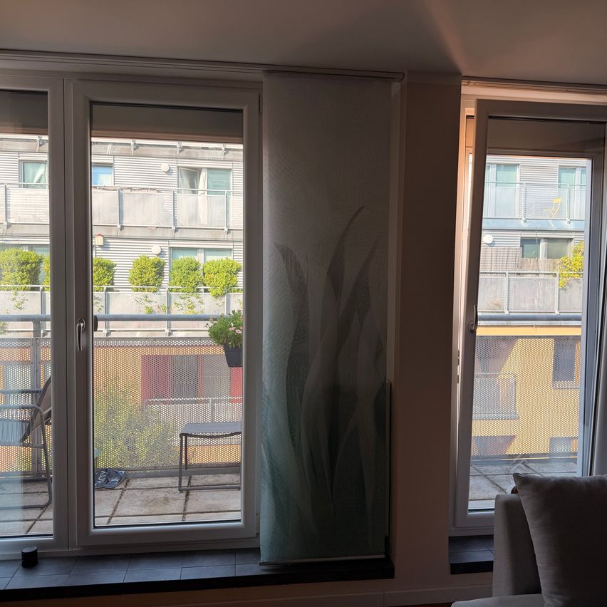 An indoor view of a balcony with a glass door, a window, and a curtain with a plant design. The balcony has a metal railing and a potted plant. Outside, there are buildings and trees.