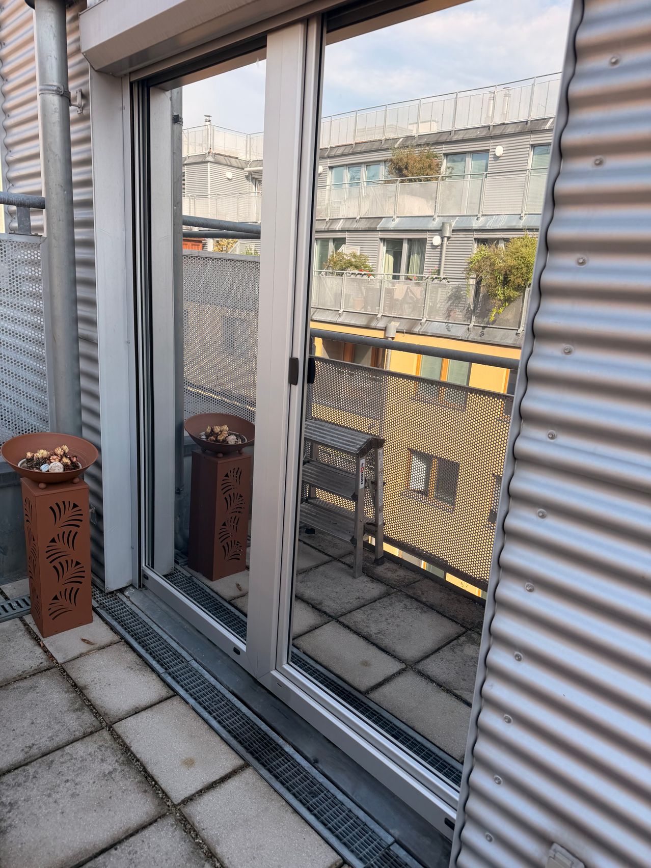 A balcony with a sliding glass door and a view of a yellow building with a balcony and a bench. There are two brown decorative bowls on the floor.