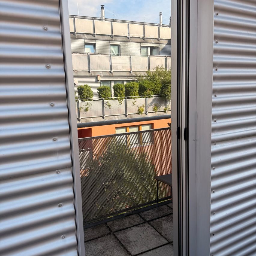 A view from a balcony showing a modern building with a balcony and a tree in the foreground. The balcony has a metal fence and a table.