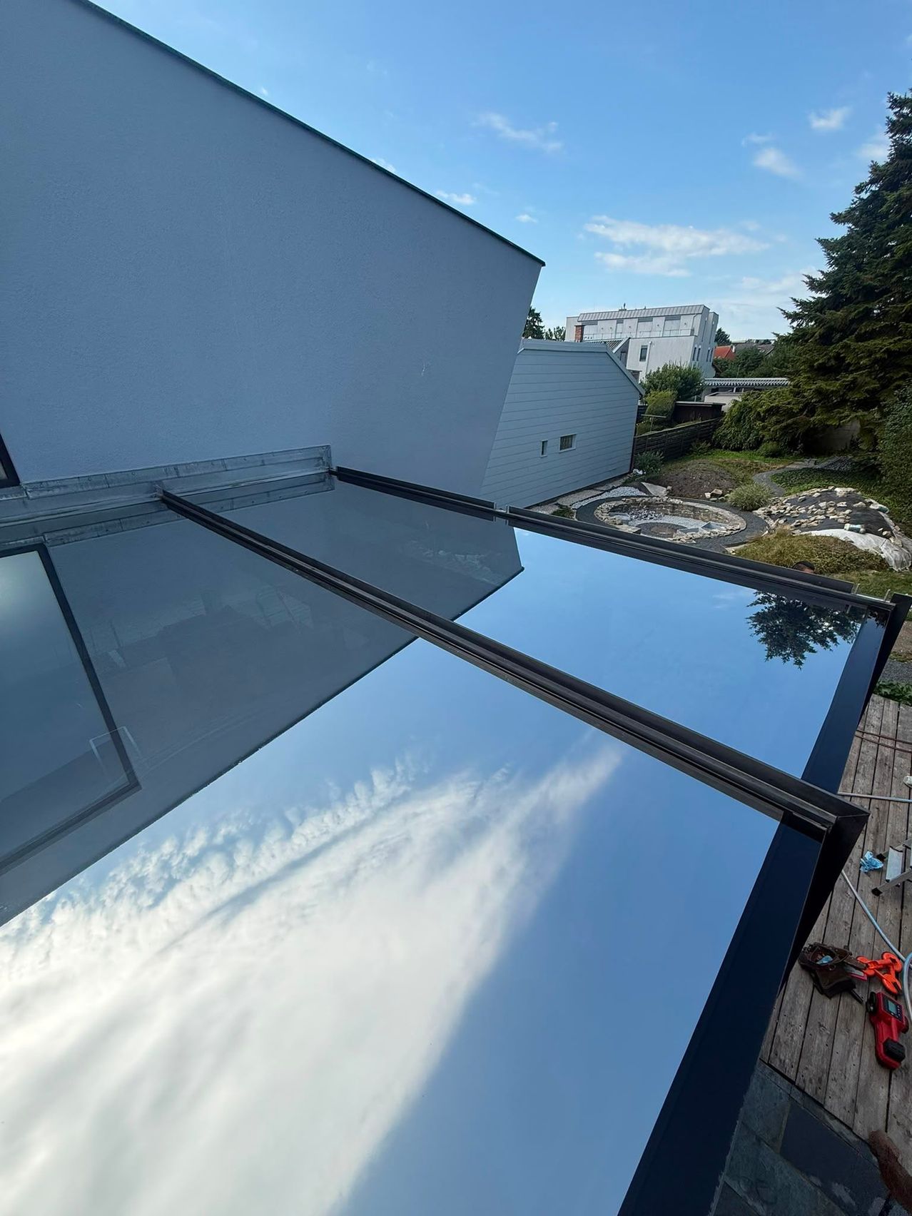 A modern glass roof with a black frame is reflected on a clear day, showing a blue sky with clouds and a house in the background.