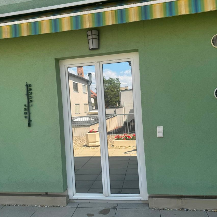 A green building exterior with a white sliding glass door, a temperature gauge, and a balcony with potted plants.