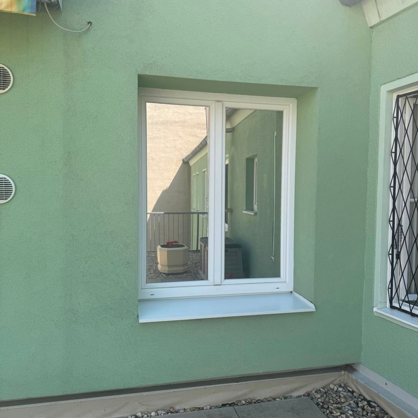 A green-painted wall with a white window frame and sill, reflecting a balcony with a potted plant and a metal railing.