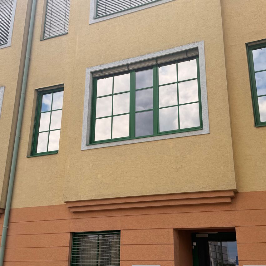 A beige building with green-framed windows and a pipe on the left side. The windows reflect the sky and clouds.