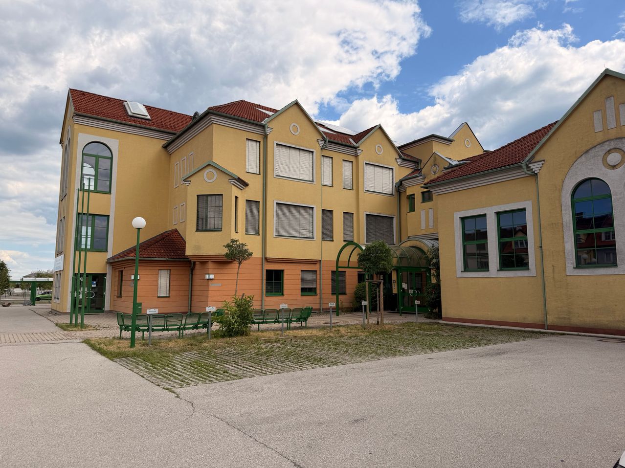 A row of yellow buildings with red roofs and green accents, surrounded by grass and benches, under a partly cloudy sky.