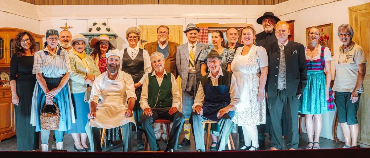 Eine Gruppe älterer Schauspieler in Vintage-Kostümen posiert auf der Bühne, einige auf Hockern sitzend. Der Hintergrund besteht aus einem roten Vorhang und einer Holzwand.