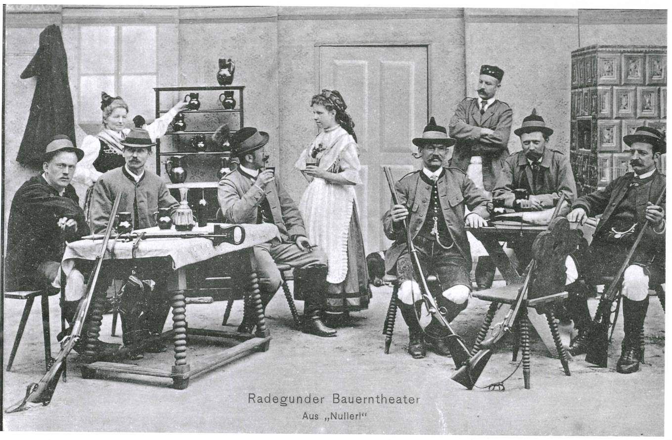Schwarz-Weiß-Foto von Menschen in historischen bayerischen Kostümen, die um einen Tisch herum sitzen. Eine Frau steht in der Mitte. Das Foto trägt die Aufschrift Radegunder Bauerntheater Aus Nuller.
