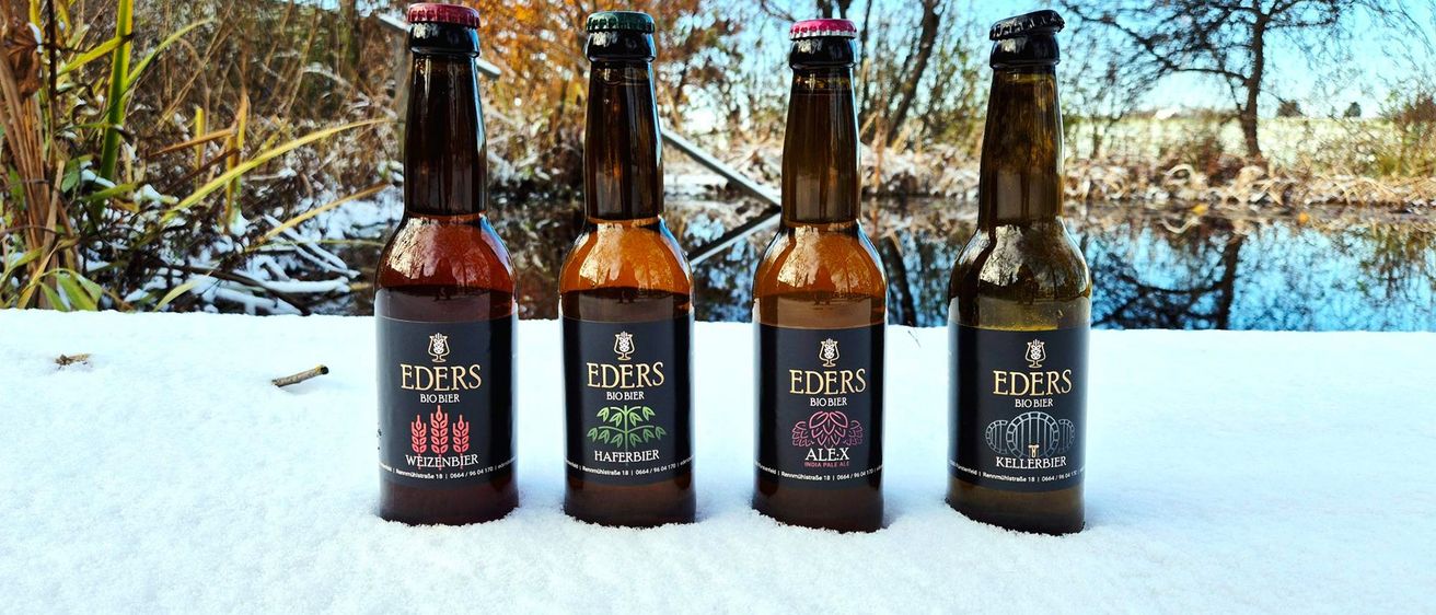 Vier Flaschen Eders Biobier stehen auf einer verschneiten Oberfläche. Drei Flaschen sind mit verschiedenen Geschmacksrichtungen beschriftet: Weizenbier, Haferbier und Alex. Die vierte Flasche ist mit Kellerbier beschriftet. Bäume stehen hinter den Flaschen.