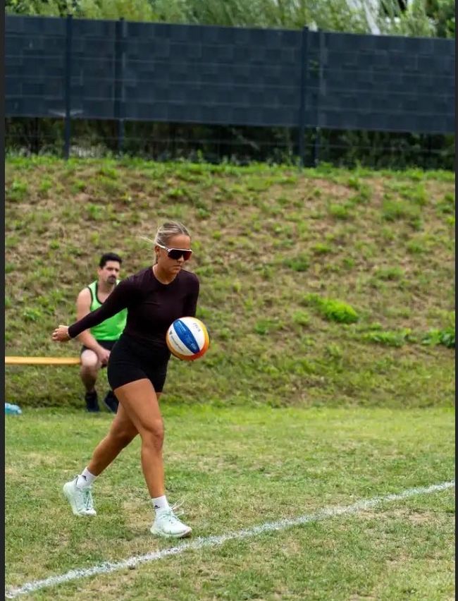 Eine Frau in einem schwarzen Outfit spielt Beachvolleyball auf einem Grasfeld. Ein Mann sitzt auf einer Bank hinter ihr.