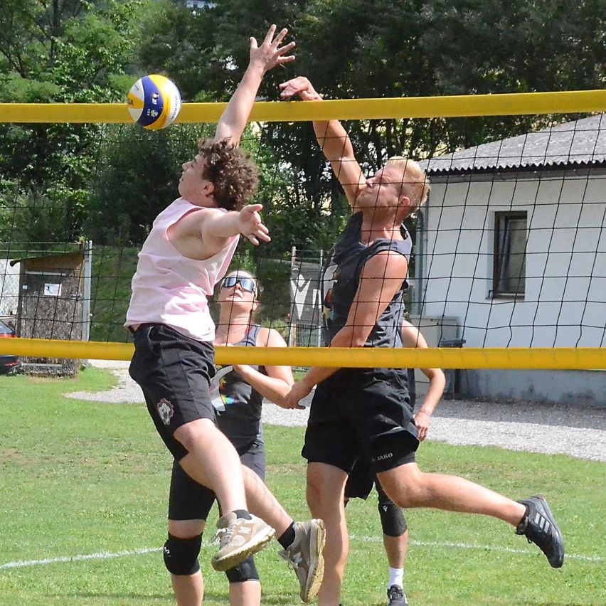 Zwei Männer spielen Volleyball auf einem Rasenfeld, einer springt, um den Ball zu blockieren, während der andere versucht, ihn über das Netz zu schlagen.