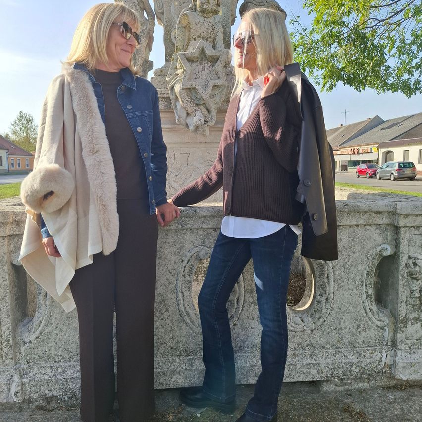 Zwei Frauen stehen zusammen, eine trägt einen Pelzmantel und die andere einen braunen Cardigan, beide halten sich an den Händen vor einer Steinmauer.