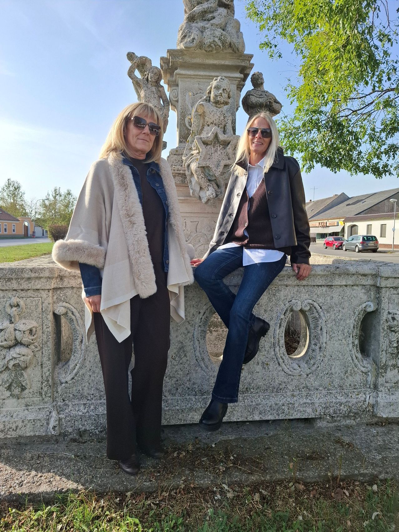 Zwei Frauen in Sonnenbrillen posieren vor einer Steinmauer mit einer Statue dahinter.