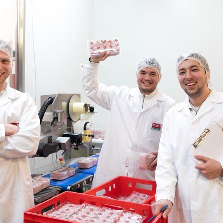 Drei Männer in Laborkittel stehen in einer Fabrik, lächeln und halten eine Packung Lebensmittel. Der Mann in der Mitte hält die Packung hoch, während der Mann auf der rechten Seite ein Klemmbrett hält. Ein Förderband mit Lebensmittelprodukten und Fabrikausrüstung ist im Hintergrund sichtbar.
