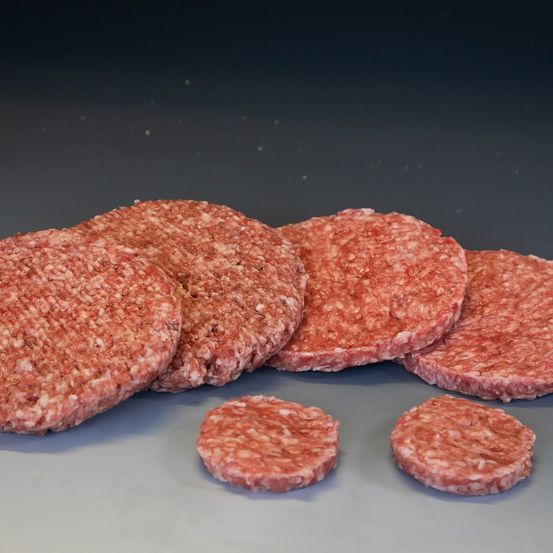 Mehrere rohe Hamburger-Patties sind auf einer dunklen Oberfläche angeordnet. Die Patties variieren in der Größe, wobei einige größer und andere kleiner sind. Sie sind roh und sehen frisch aus.