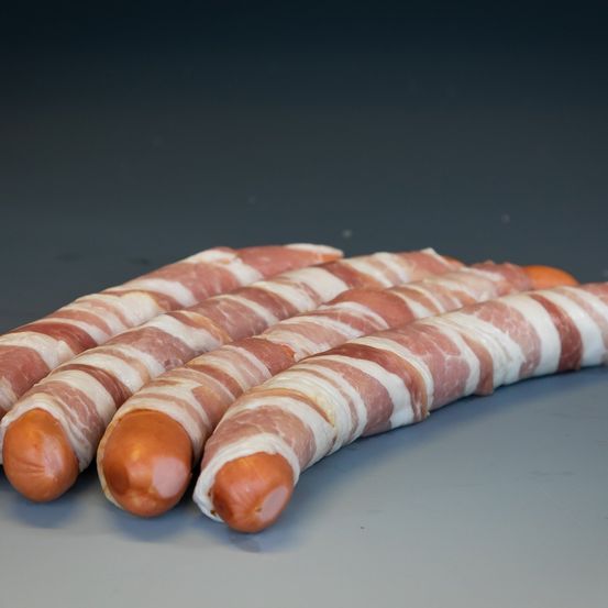 Vier Würste sind in Speck eingewickelt, wobei jede Scheibe leicht überlappt. Sie liegen auf einer dunklen, flachen Oberfläche.