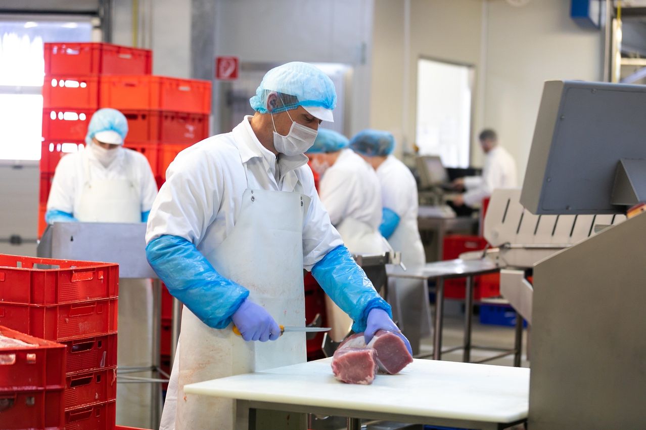 Ein Mann in einem Fleischverarbeitungsbetrieb trägt einen weißen Mantel, eine Maske und Handschuhe und schneidet Fleisch. Hinter ihm sind andere Arbeiter anwesend, und Lagerboxen sind gestapelt.