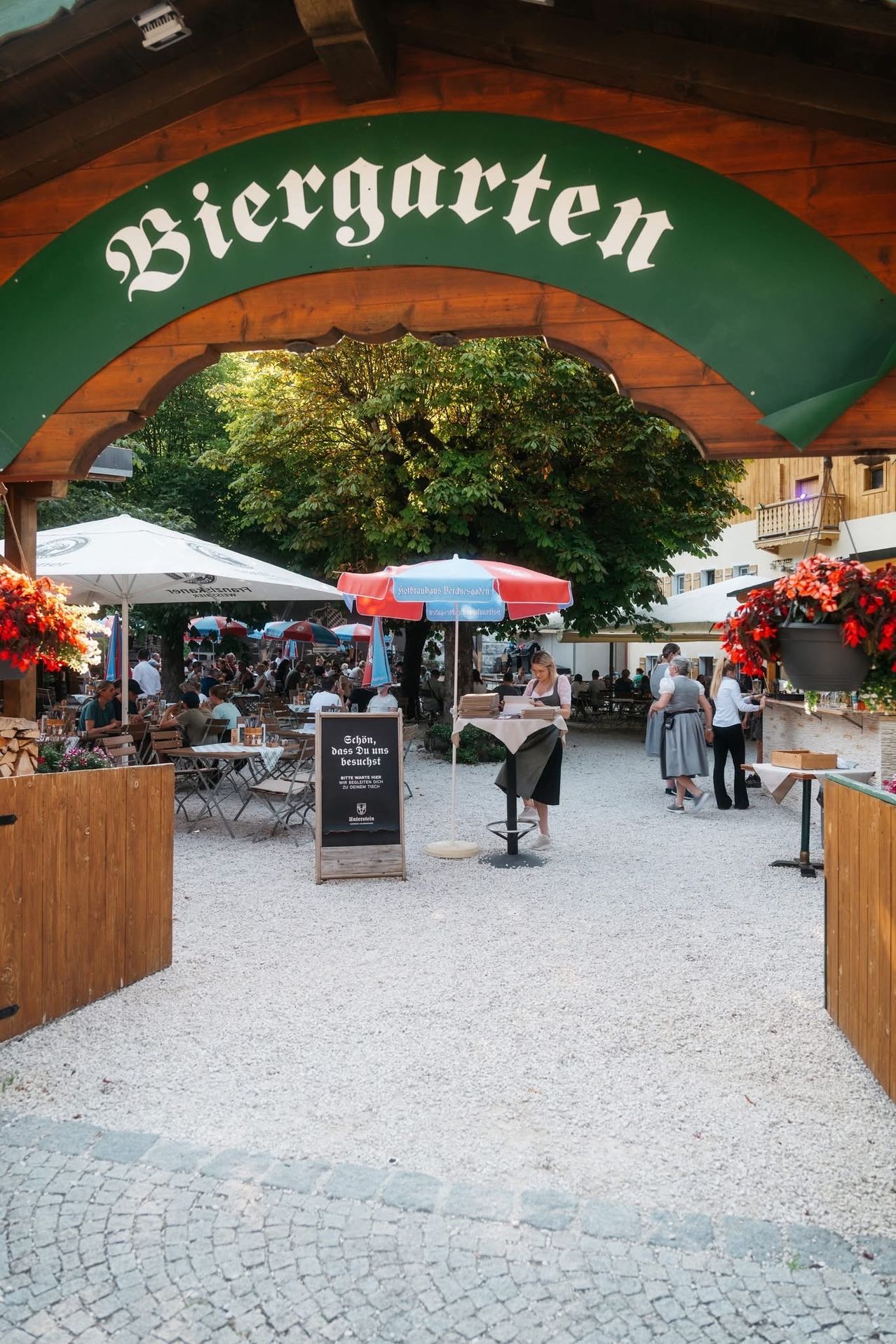 A bustling outdoor dining area with tables, chairs, and people. The entrance is marked with a green sign. Trees and plants add to the ambiance. A woman stands behind a table with a menu.