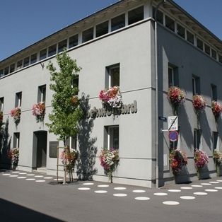 Ein graues Gebäude mit mehreren Fenstern hat Blumenvasen an der Außenwand. Es hat ein Schild mit dem Wort 'Floral' darauf. Vor dem Gebäude stehen Bäume und Pflanzen.