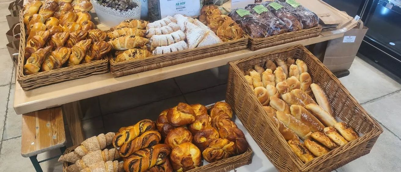 Ein Bäckerei-Ausstellungstisch mit verschiedenen Gebäcksorten in Weidenkörben, darunter Croissants, Brötchen und süßes Gebäck. Im Hintergrund befinden sich Weinflaschen, eine Topfpflanze und ein Schild.