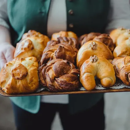 Eine Person hält eine Tablett mit verschiedenen Gebäckstücken, darunter Croissants, Brötchen und Brötchen, alle mit sichtbaren Walnüssen darauf. Das Tablett liegt auf einem Spitzendeckchen.