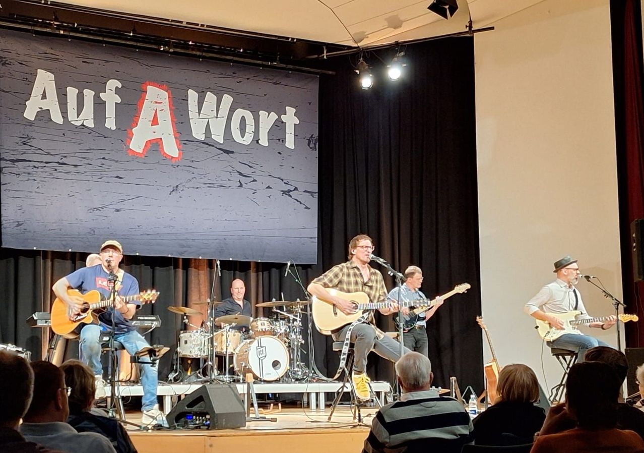 Eine Band tritt auf einer Bühne mit einem Banner auf, das 'f A Wort' steht. Die Bandmitglieder spielen Gitarren, Trommeln und Becken. Ein Mann in der Mitte sitzt und spielt Gitarre. Das Publikum sitzt vor ihnen.