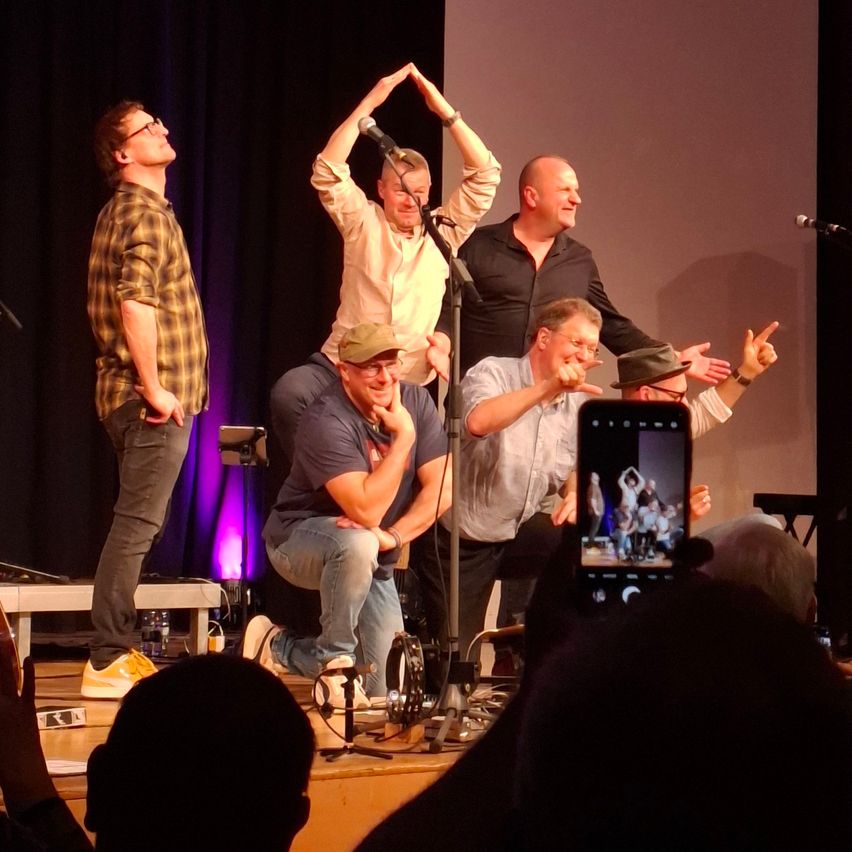 Eine Gruppe von Menschen steht auf der Bühne und führt eine Comedy-Szene auf. Eine Person kniet und eine andere hält ein Mikrofon. Zuschauer machen Fotos.