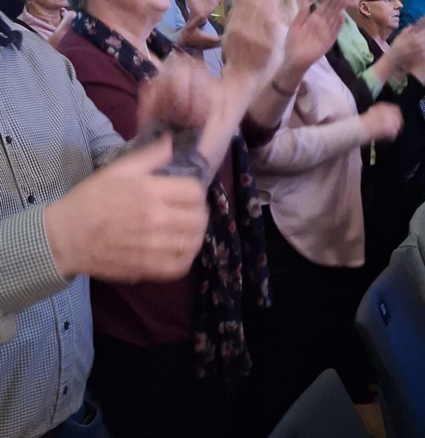 Eine Gruppe von Menschen steht und klatscht bei einer Veranstaltung, möglicherweise einer Konferenz. Eine Person trägt einen Ring.