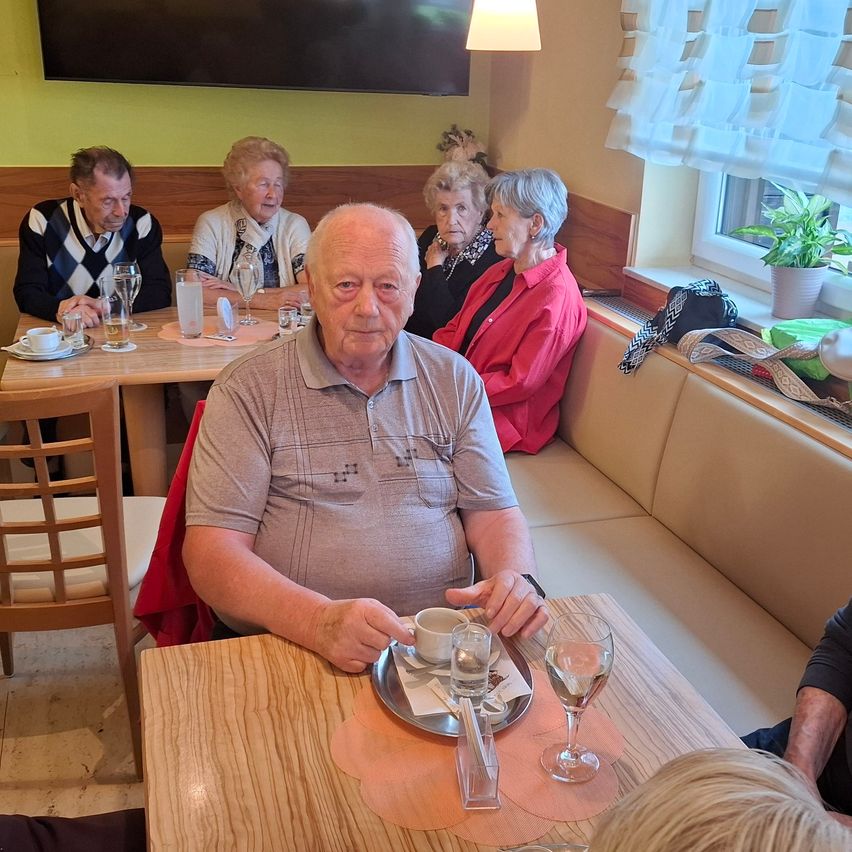 Eine Gruppe älterer Menschen sitzt in einem Café. Ein Mann sitzt an einem Tisch und trinkt Kaffee. Auf dem Tisch stehen Weingläser. Der Raum hat ein Fenster mit einer Topfpflanze.